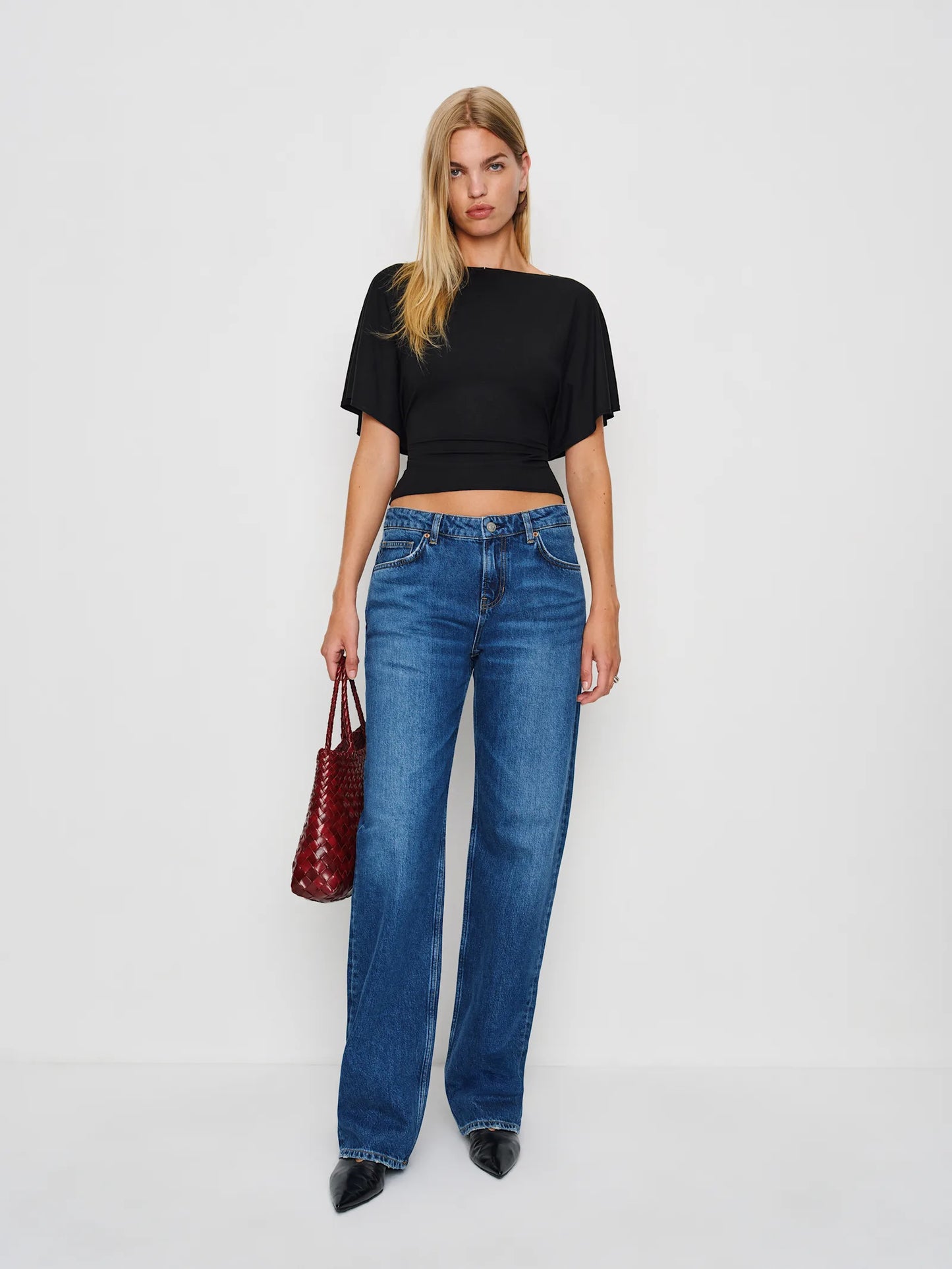 Woman wearing blue jeans and a black top, holding a red handbag on a white background