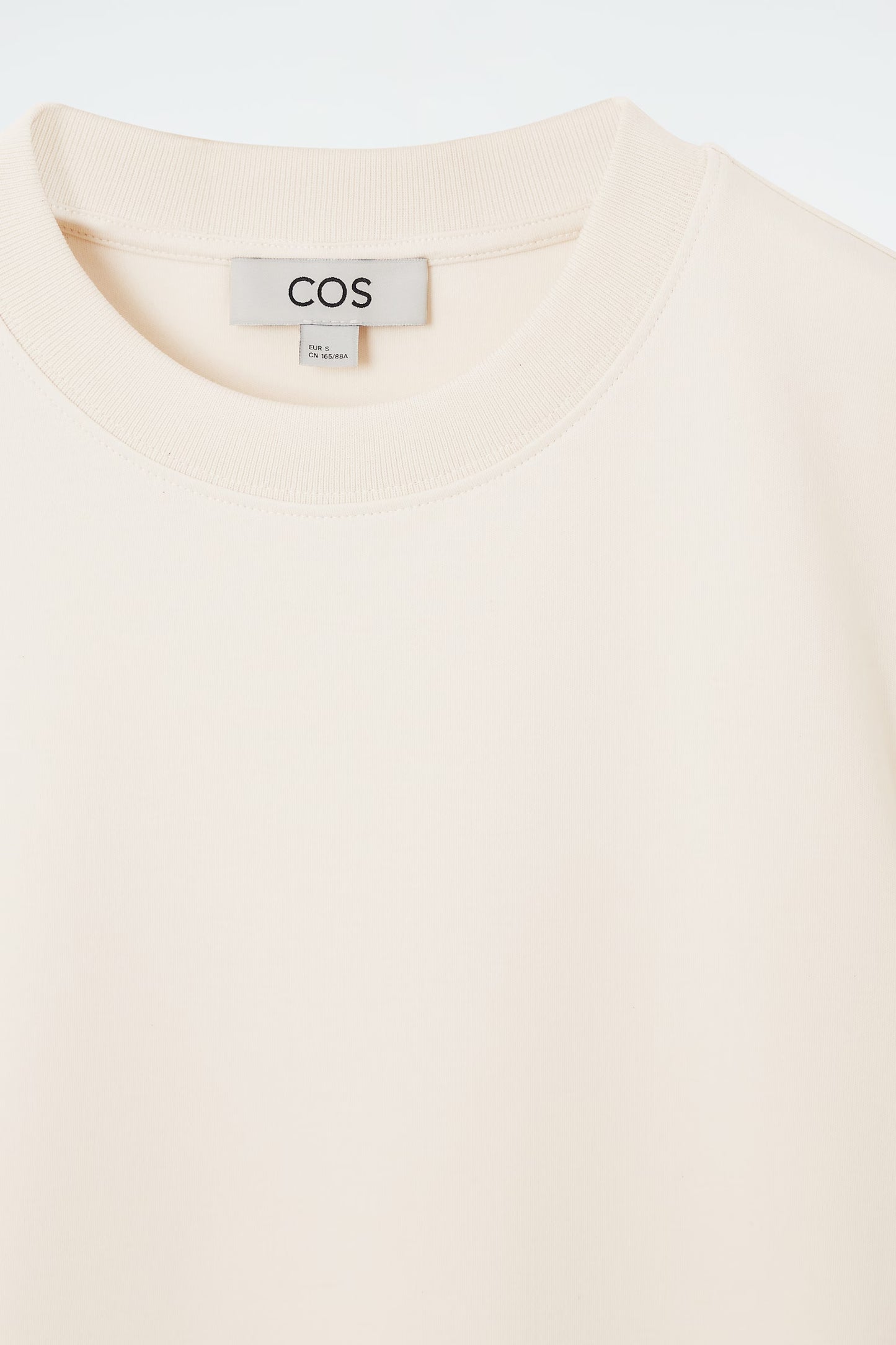 Close-up of a cream-colored garment with a COS label on a light gray background