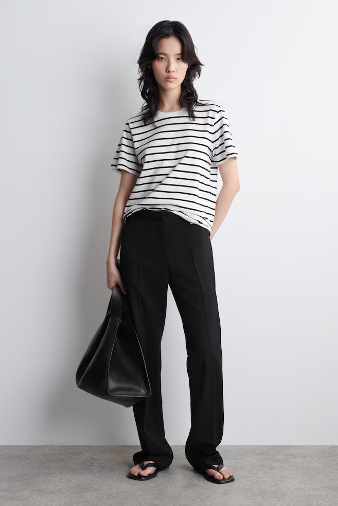 Woman wearing a black and white striped shirt and black pants, holding a black bag against a plain background.