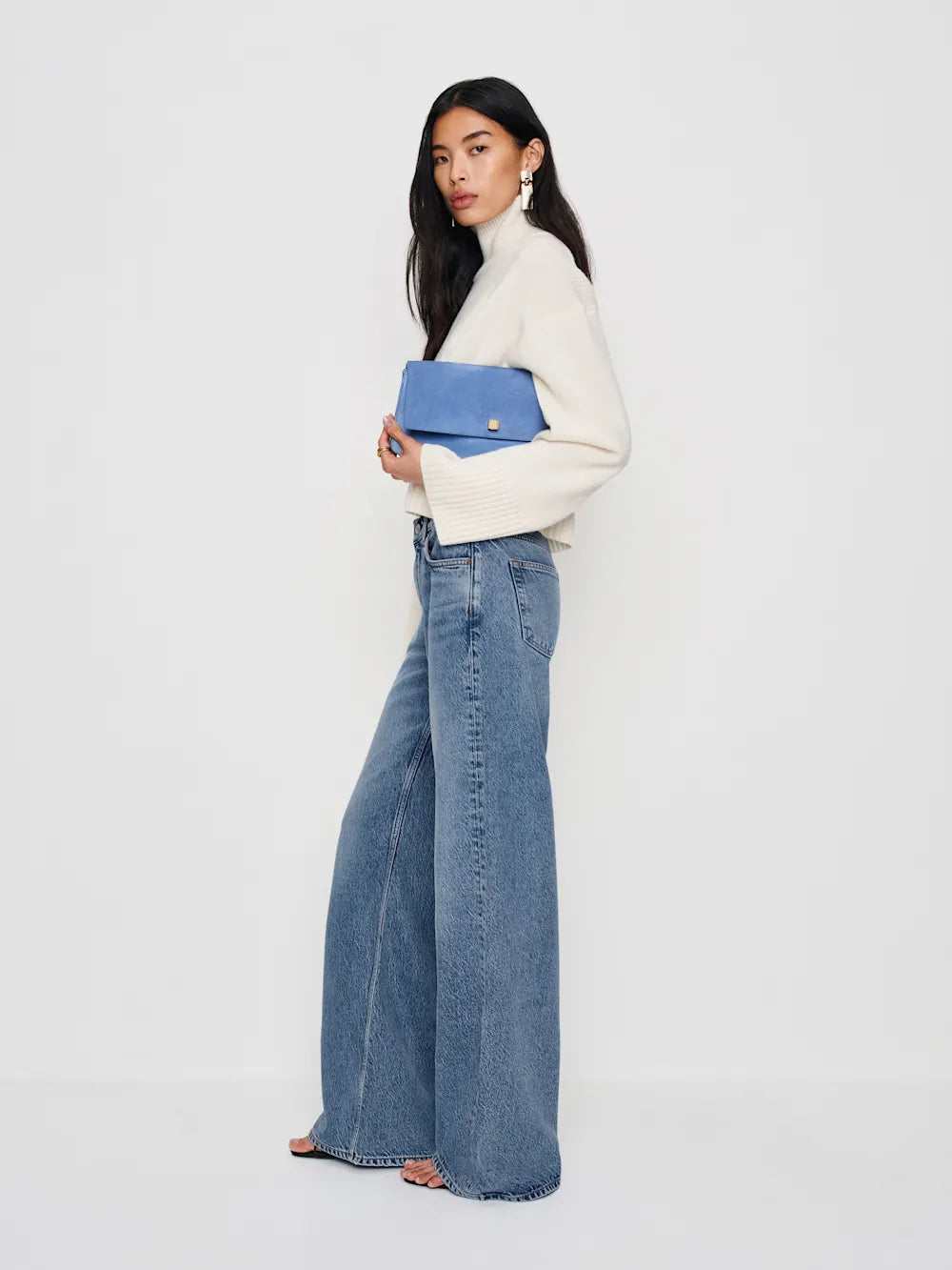 Woman wearing a white turtleneck and blue jeans holding a blue clutch against a white background