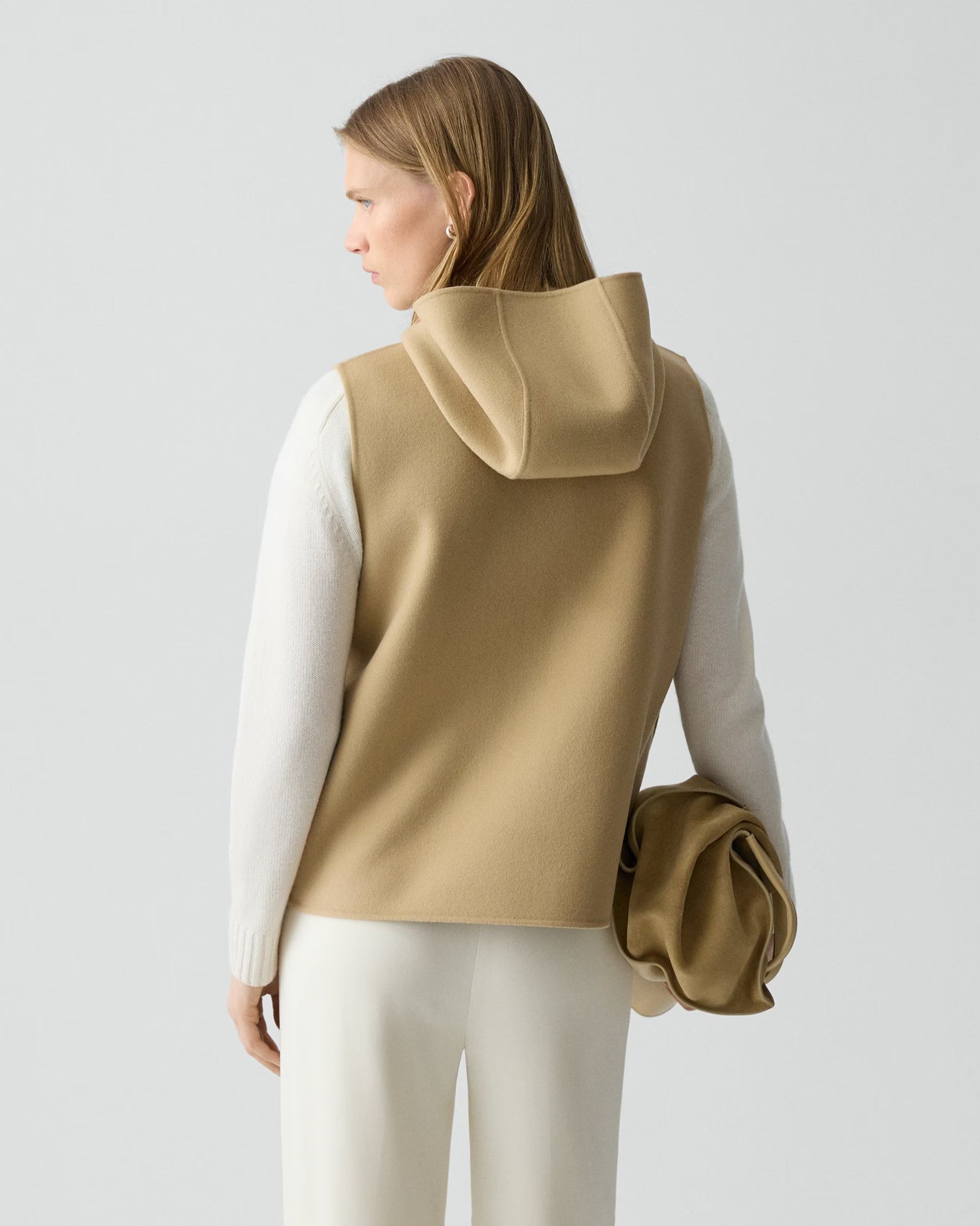 Woman wearing a beige hooded vest over a white long-sleeve shirt on a light gray background