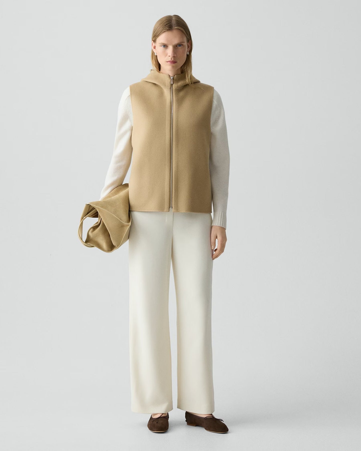 Woman wearing a beige vest over a white shirt with white pants on a light gray background