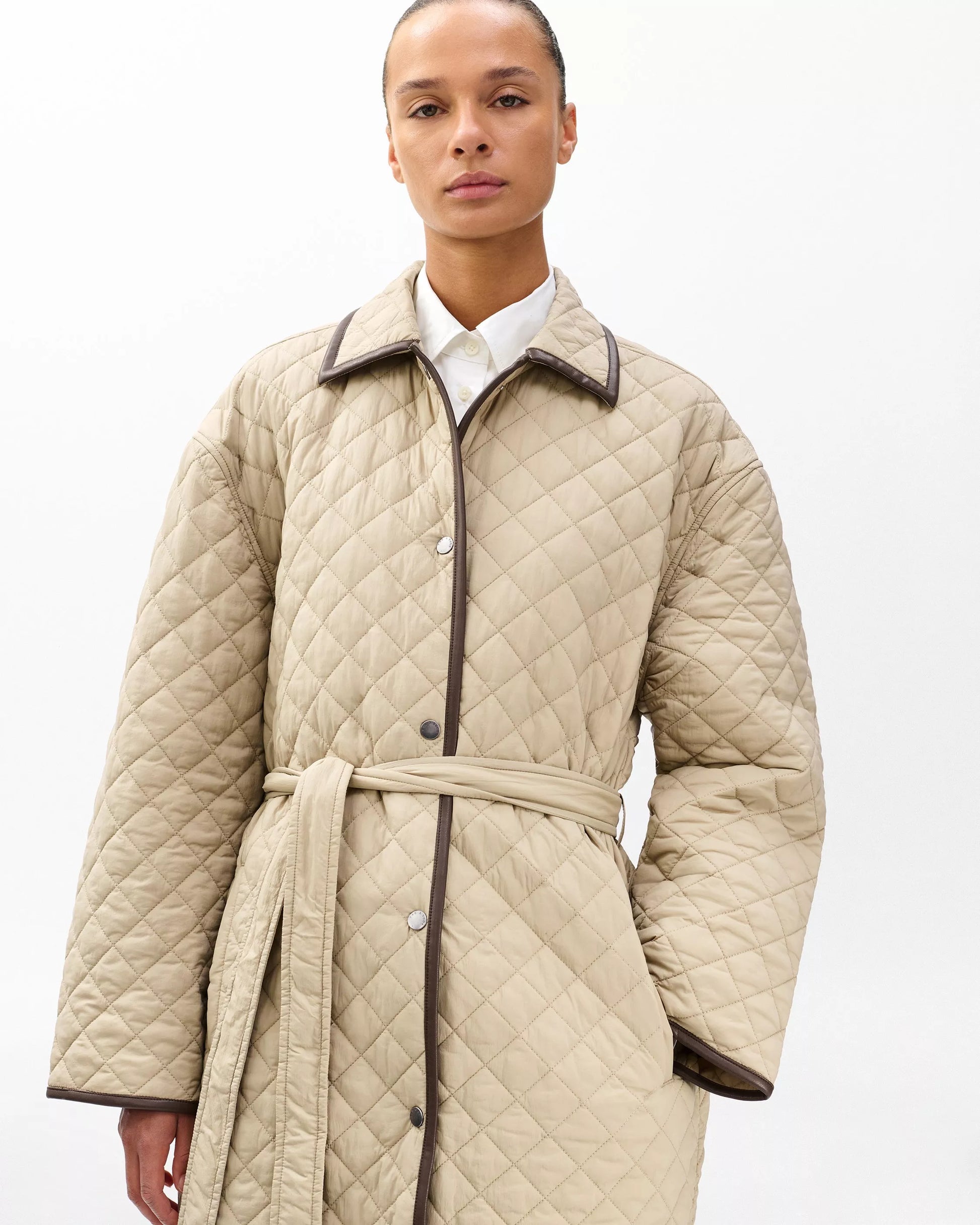 Woman wearing a beige quilted coat with a white shirt underneath on a white background