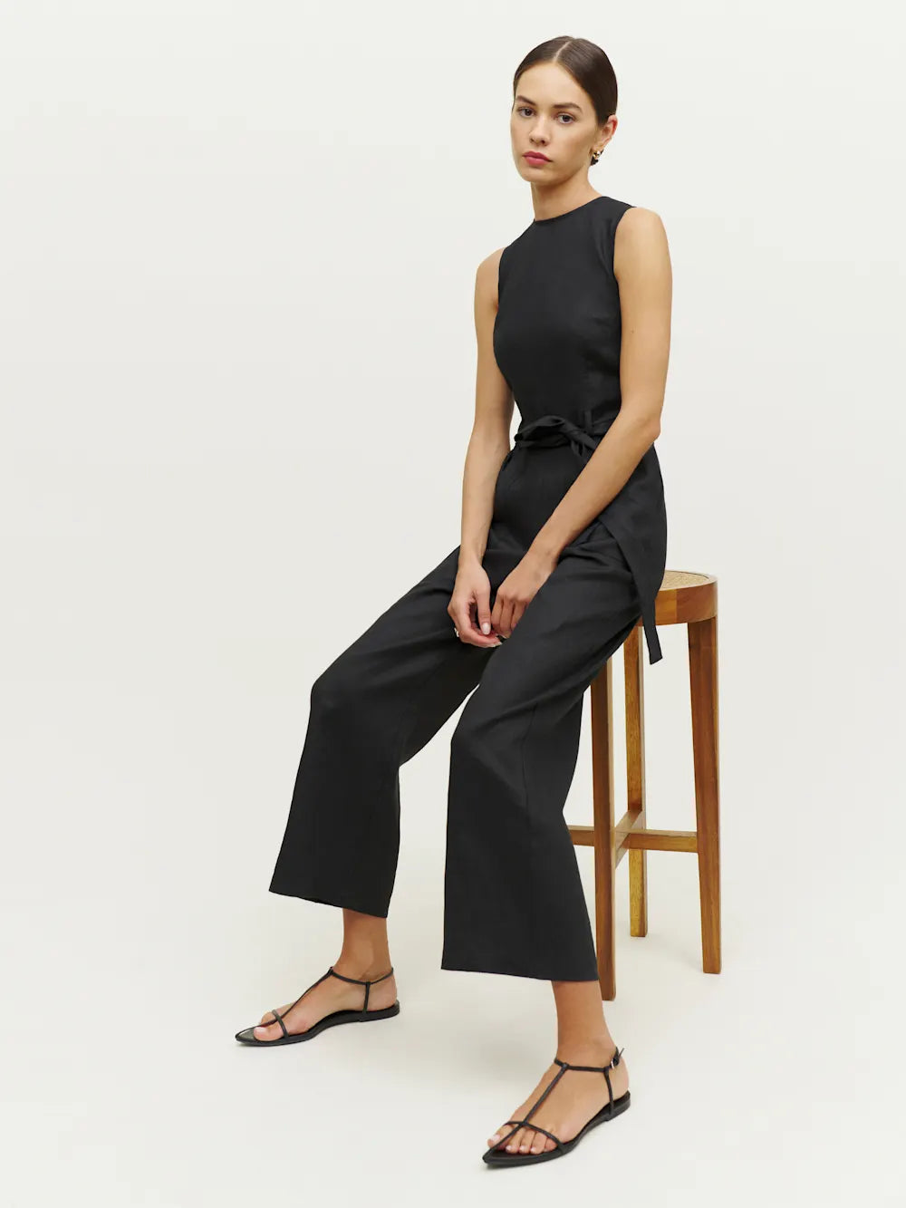 Woman wearing a black sleeveless jumpsuit sitting on a wooden stool against a white background