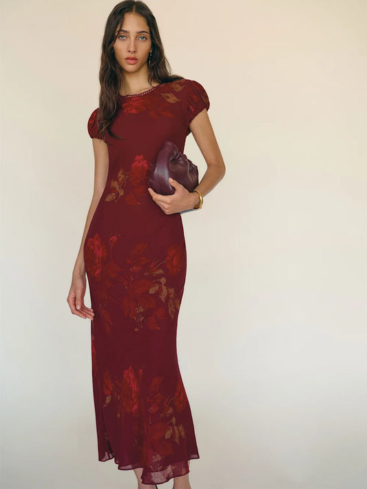 Woman wearing a burgundy floral dress holding a matching clutch against a plain background
