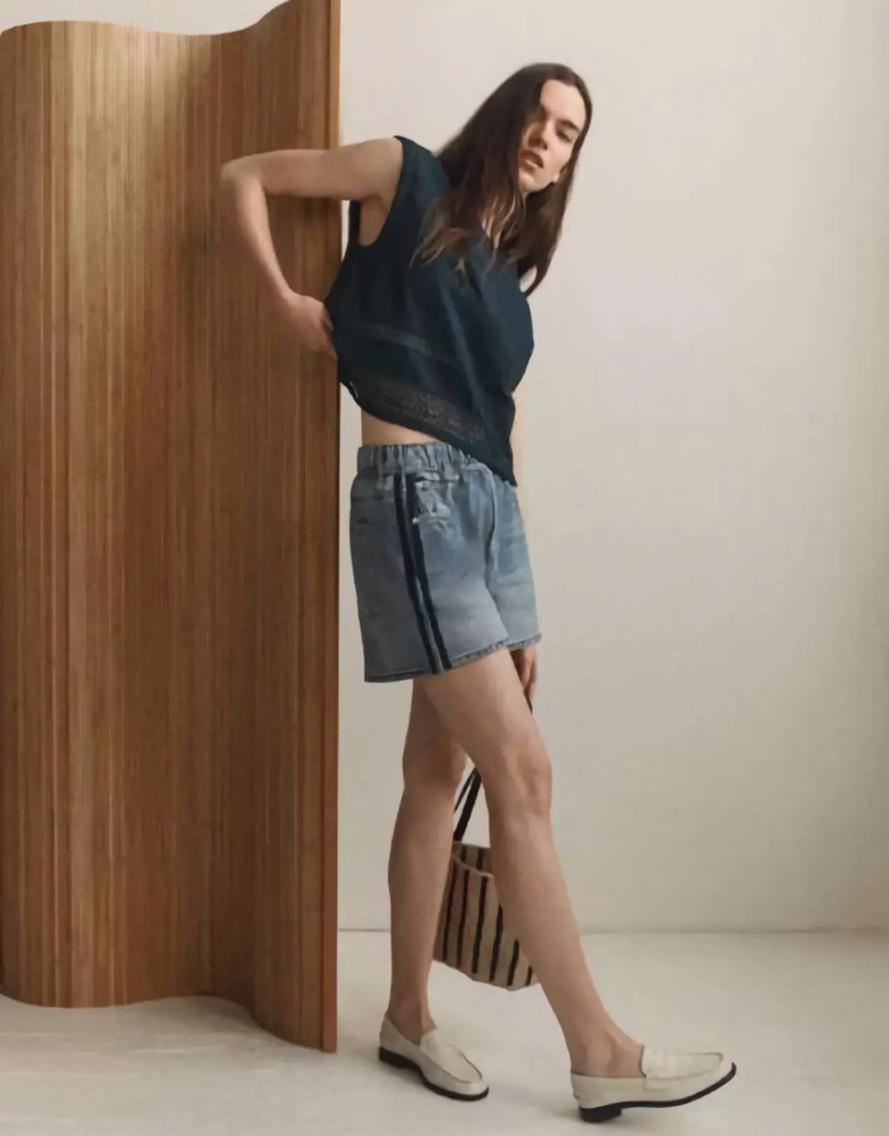 Woman in a blue top and denim shorts standing against a wooden wall.