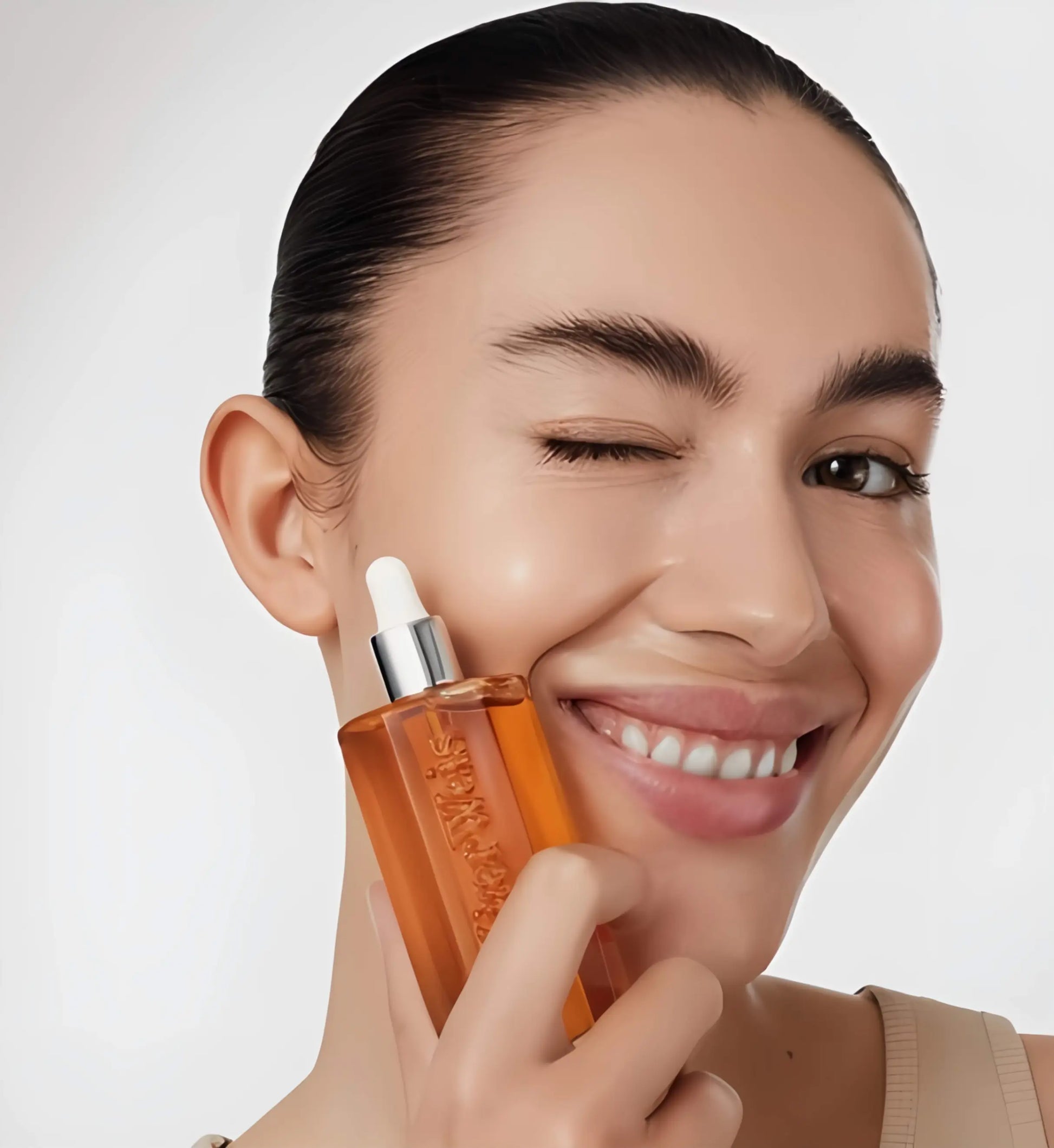 The Beautiful Oil in its iconic Kjaer Weis packaging, with a drop of the golden elixir on skin, showcasing its dewy, non-greasy finish