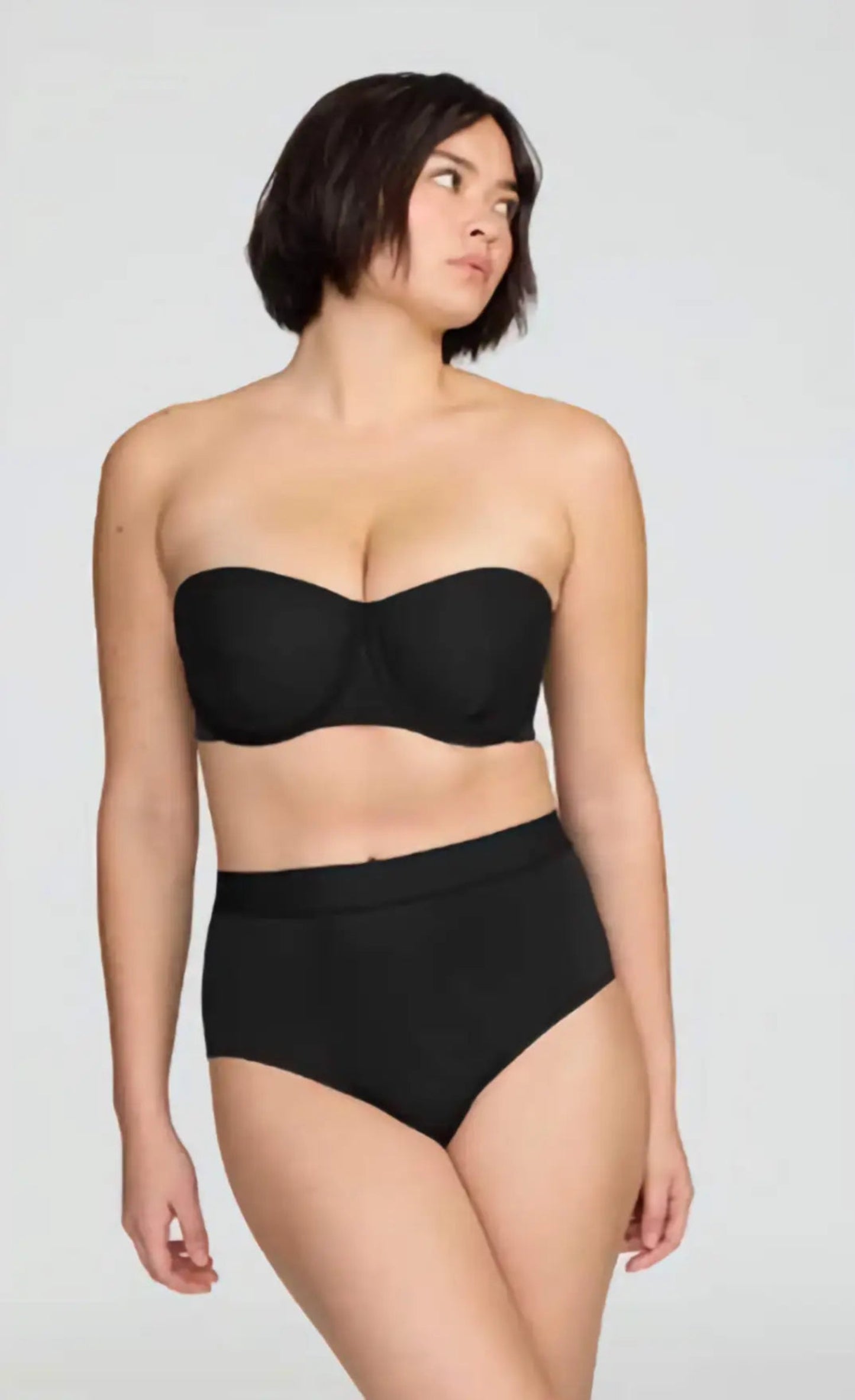 Woman wearing a black strapless top and high-waisted black underwear on a plain background